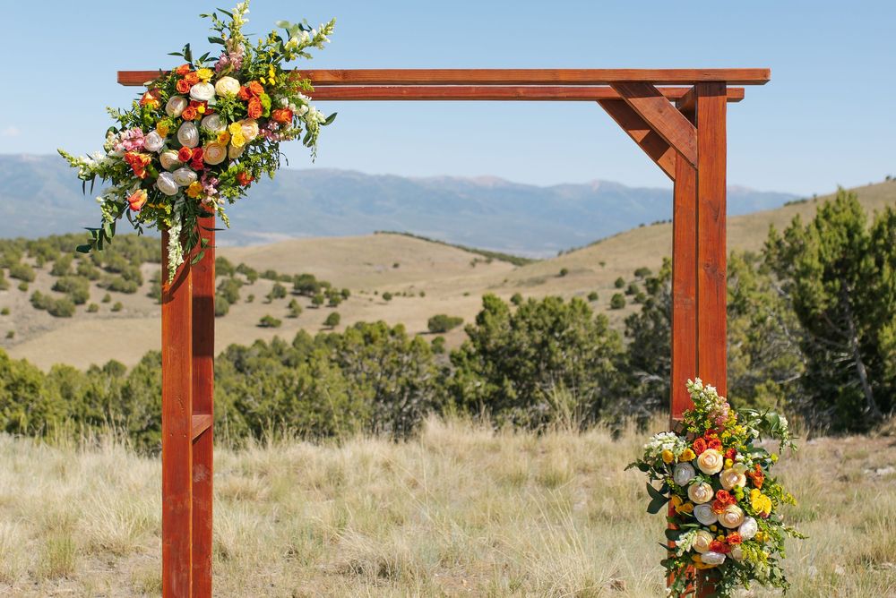 Rustic wooden wedding arch adorned with vibrant orange, yellow, and pink floral arrangements against mountain backdrop.