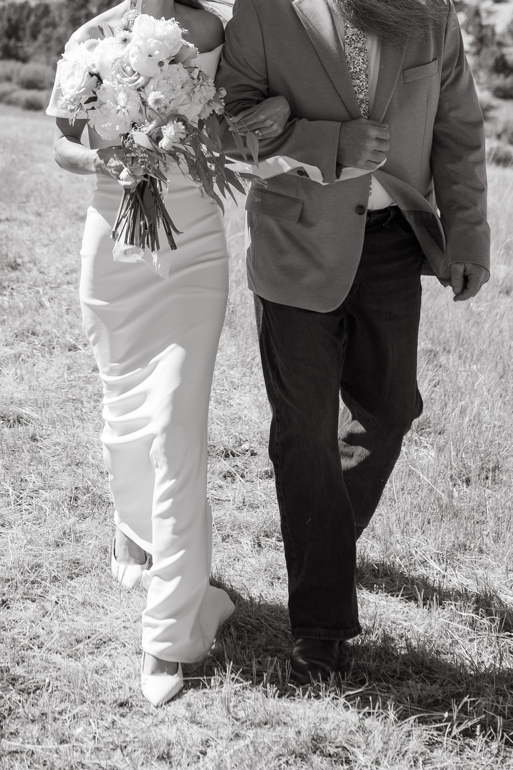 Black and white photo of bride in white pantsuit holding bouquet walking with escort through grassy field.