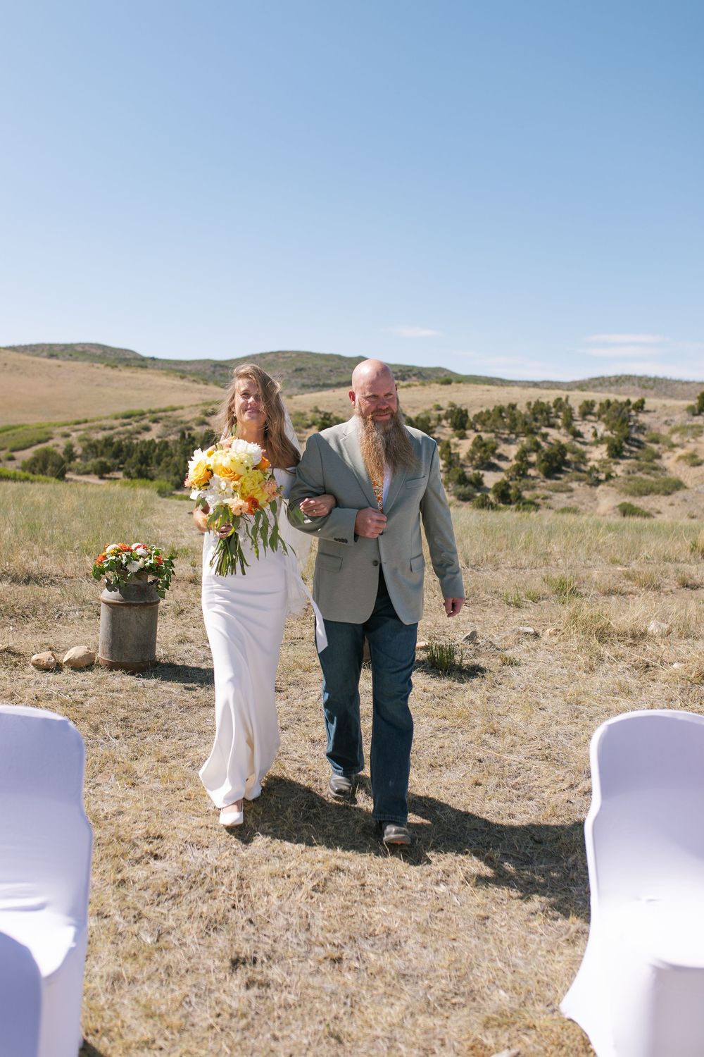 Bride in white gown walks down outdoor aisle with bearded man in gray jacket holding yellow bouquet.