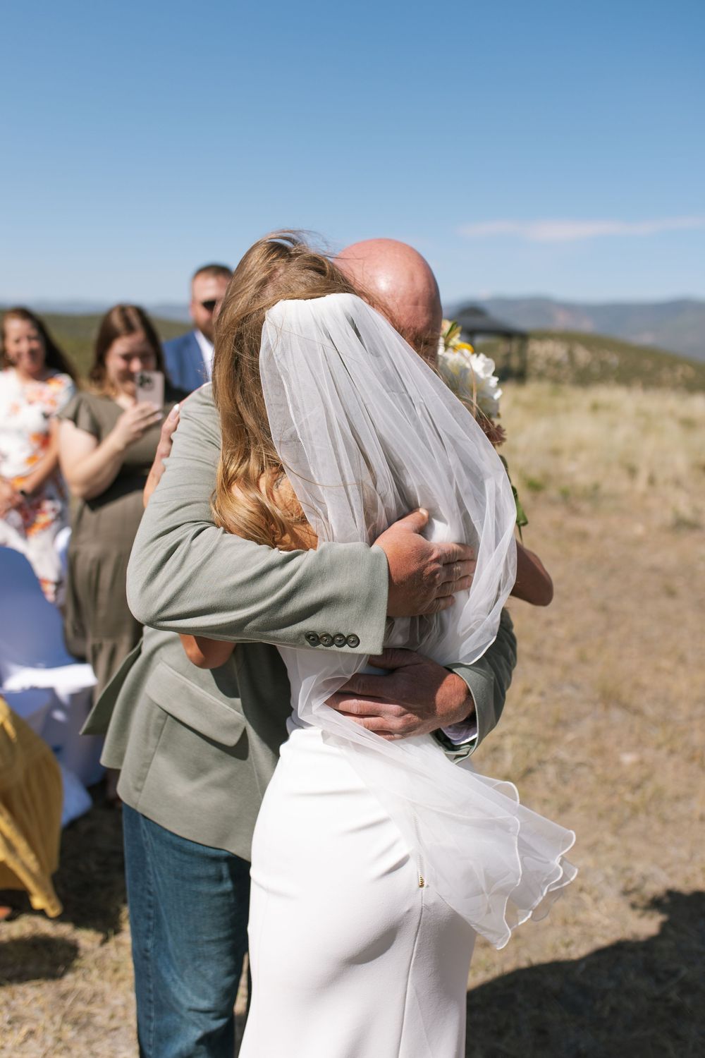 Bride in white dress and veil shares emotional hug with man in gray jacket at outdoor wedding.
