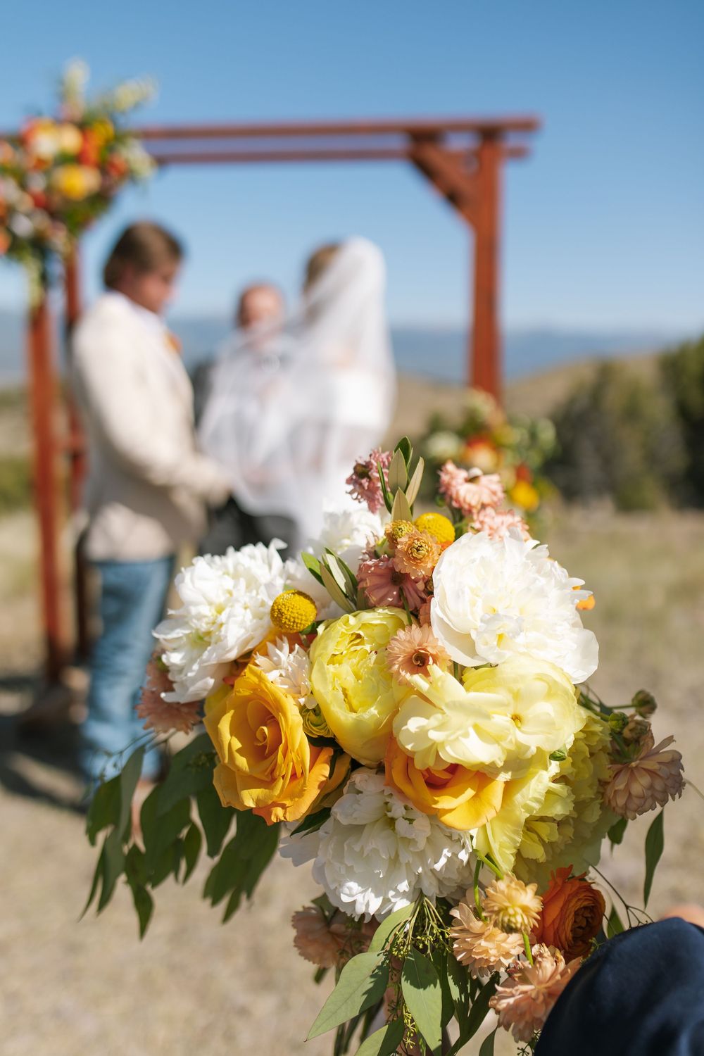 Colorful yellow and white wedding bouquet in foreground with couple exchanging vows at rustic arch.
