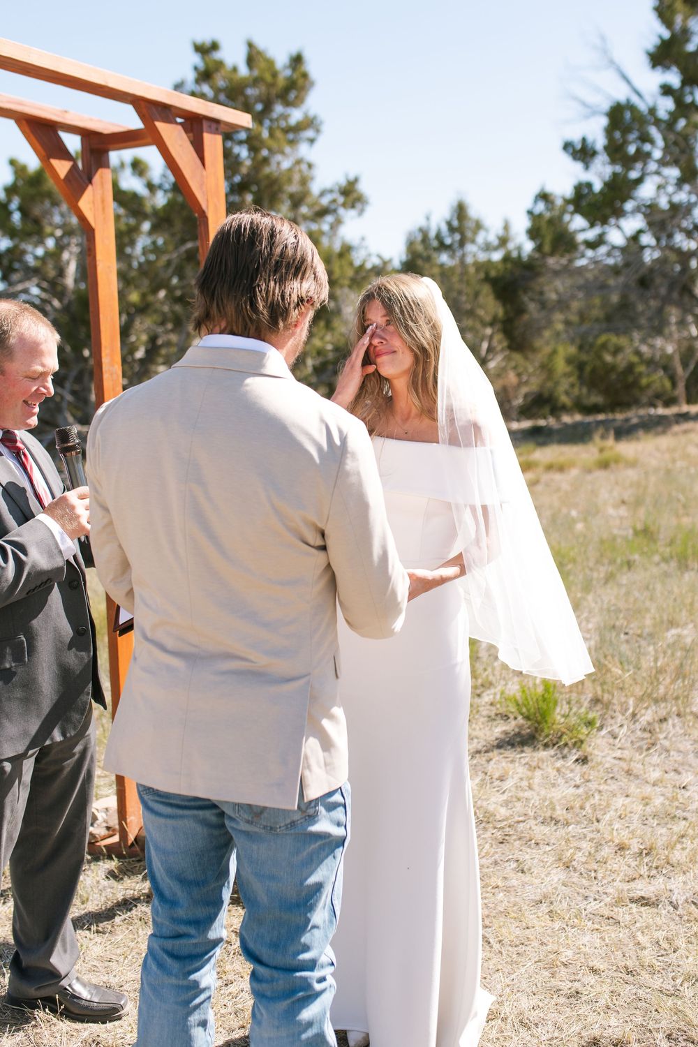 Emotional bride in white off-shoulder gown wipes tears during outdoor wedding ceremony at wooden arch.