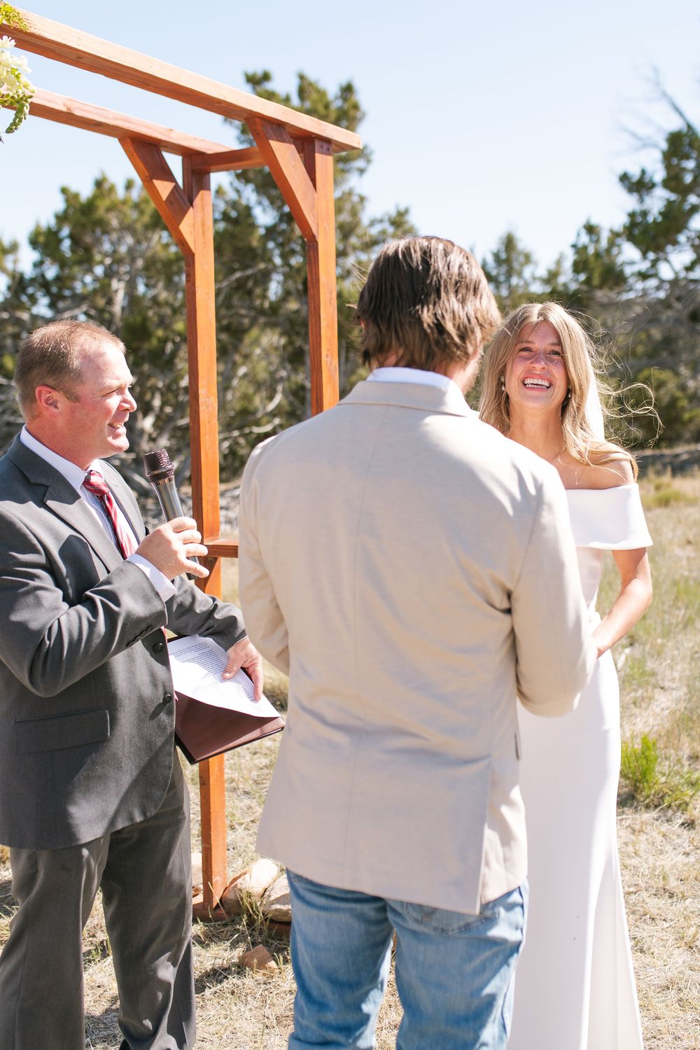 Bride laughs joyfully during outdoor wedding ceremony with groom and officiant at wooden arch.
