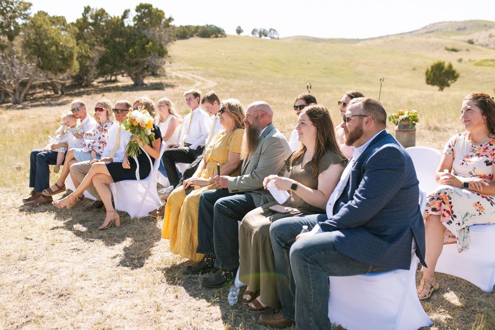Wedding guests seated in white chairs watching outdoor ceremony in scenic open field landscape.