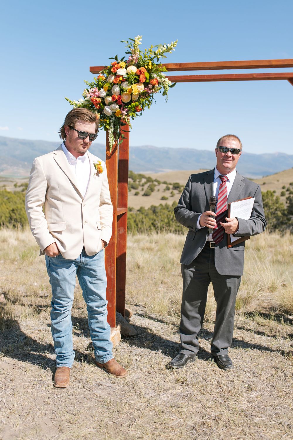 Groom in beige suit and jeans standing with officiant at floral wooden arch in mountain setting.