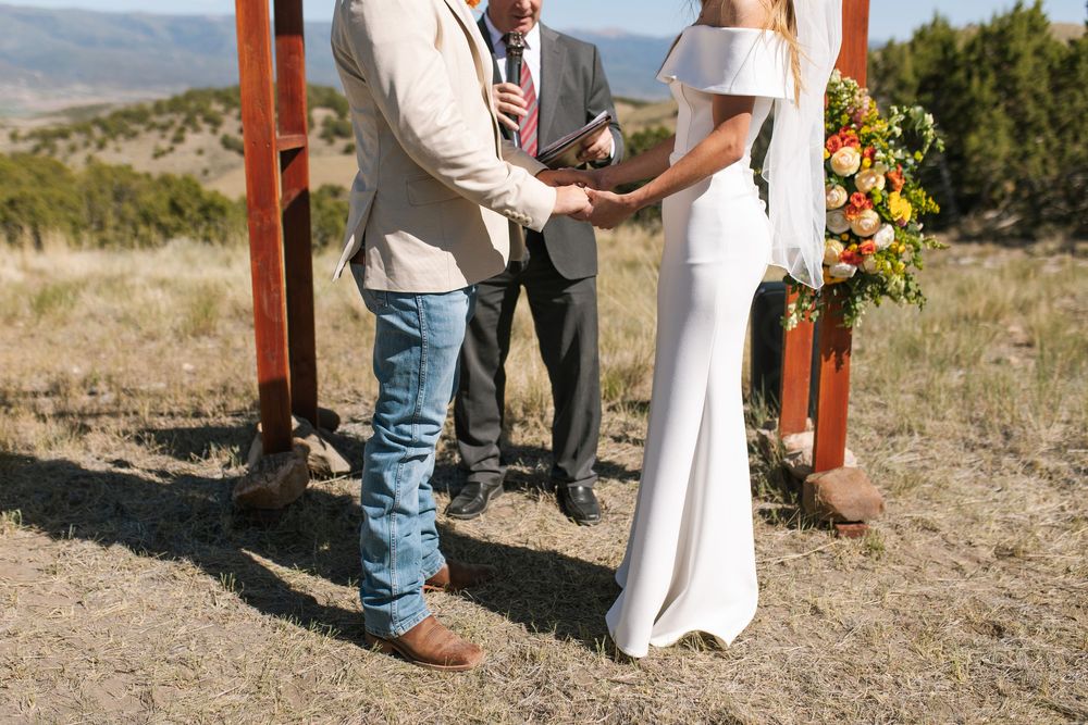 Couple holds hands during outdoor wedding ceremony at rustic wooden arch with mountain backdrop.