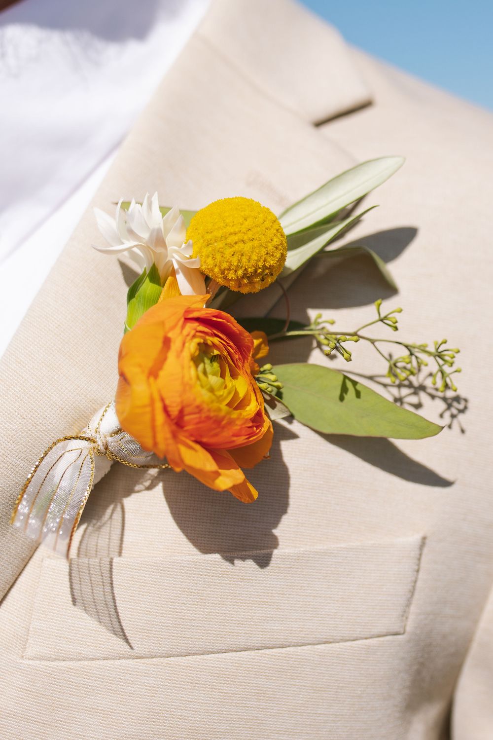 Orange ranunculus and yellow craspedia boutonniere with eucalyptus on beige groom's suit lapel.