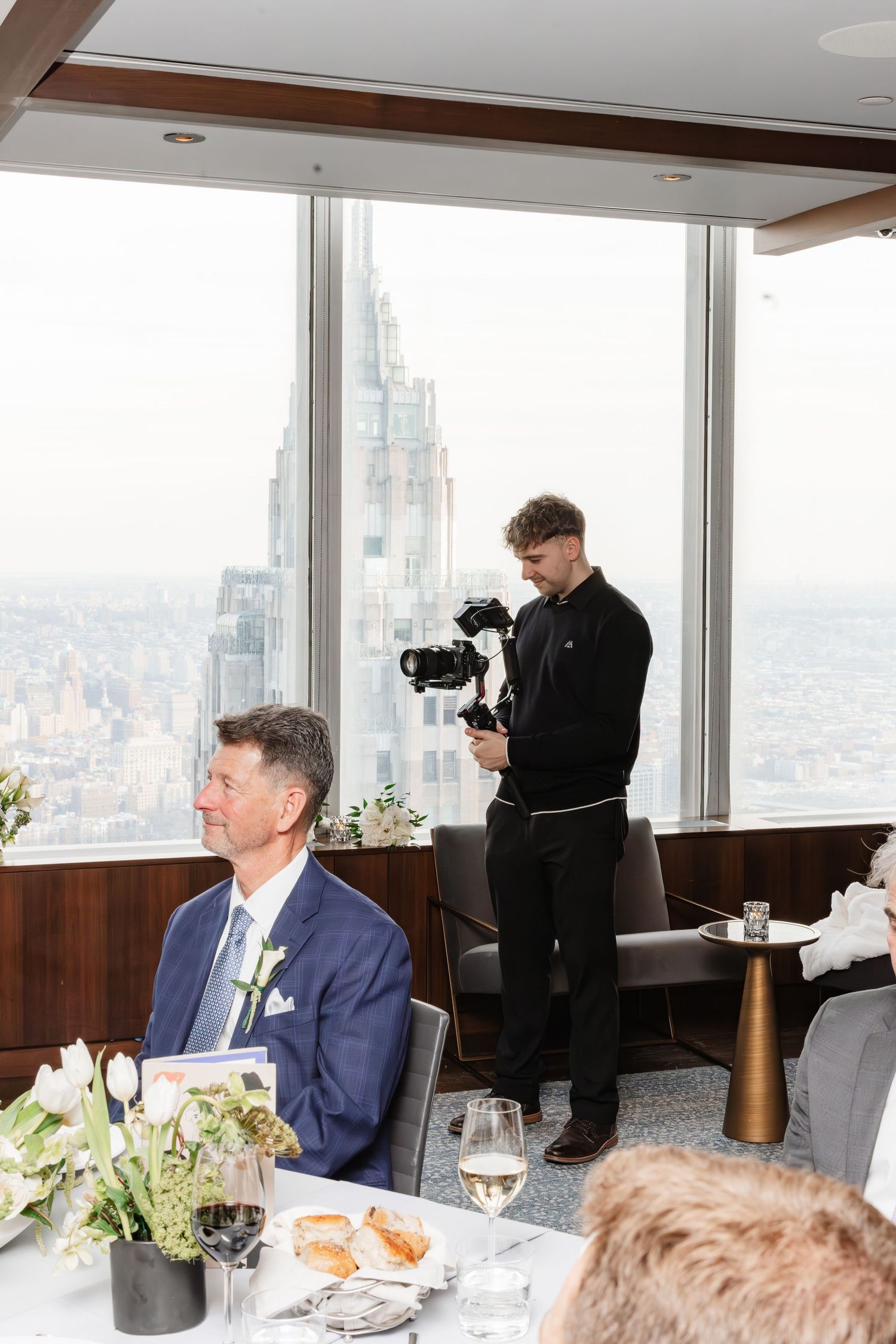 Videographer filming wedding reception guests with NYC skyline visible through floor-to-ceiling windows in high-rise venue.