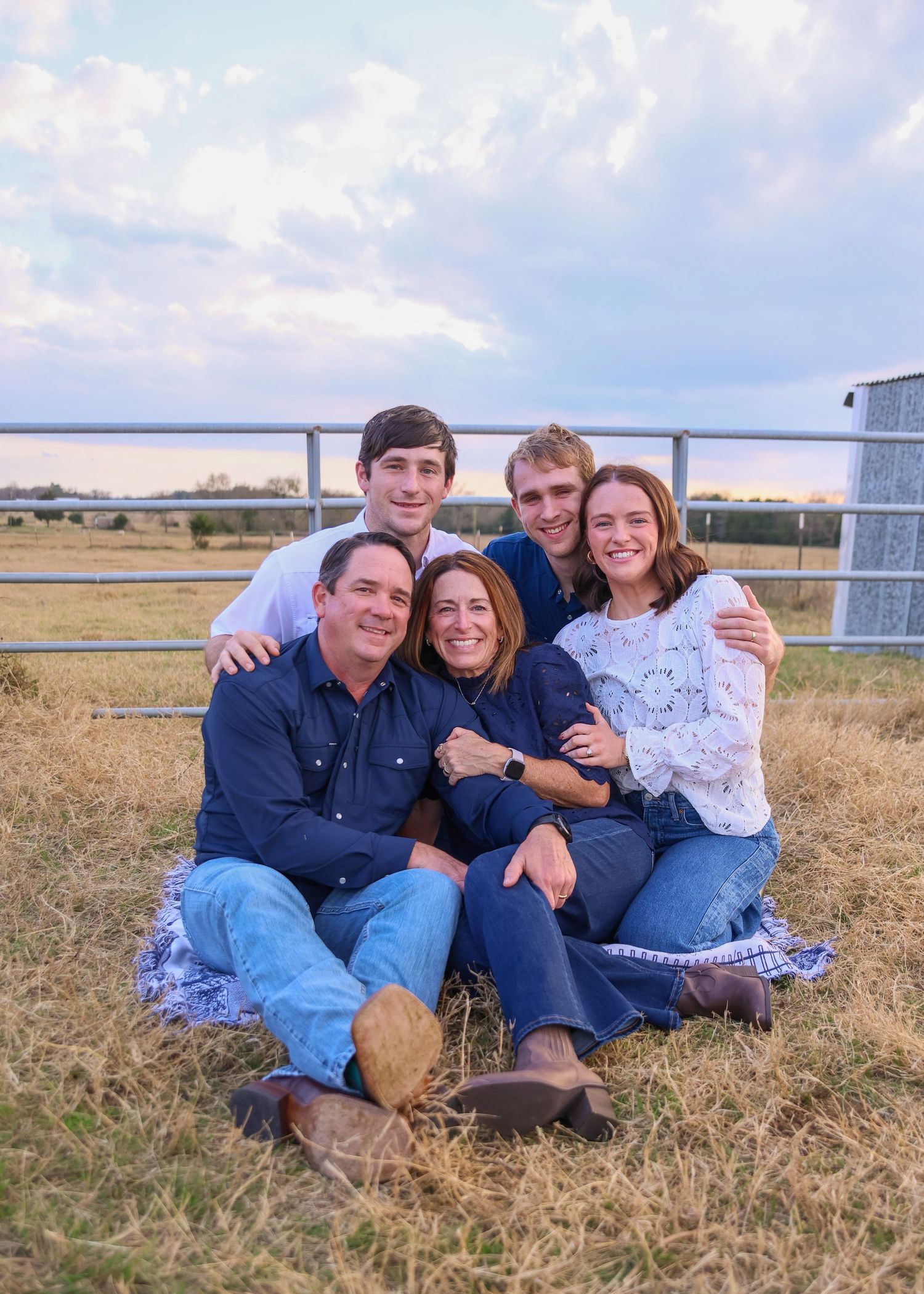 A happy family of five poses together on a grassy farm field at sunset, wearing coordinated blue outfits.