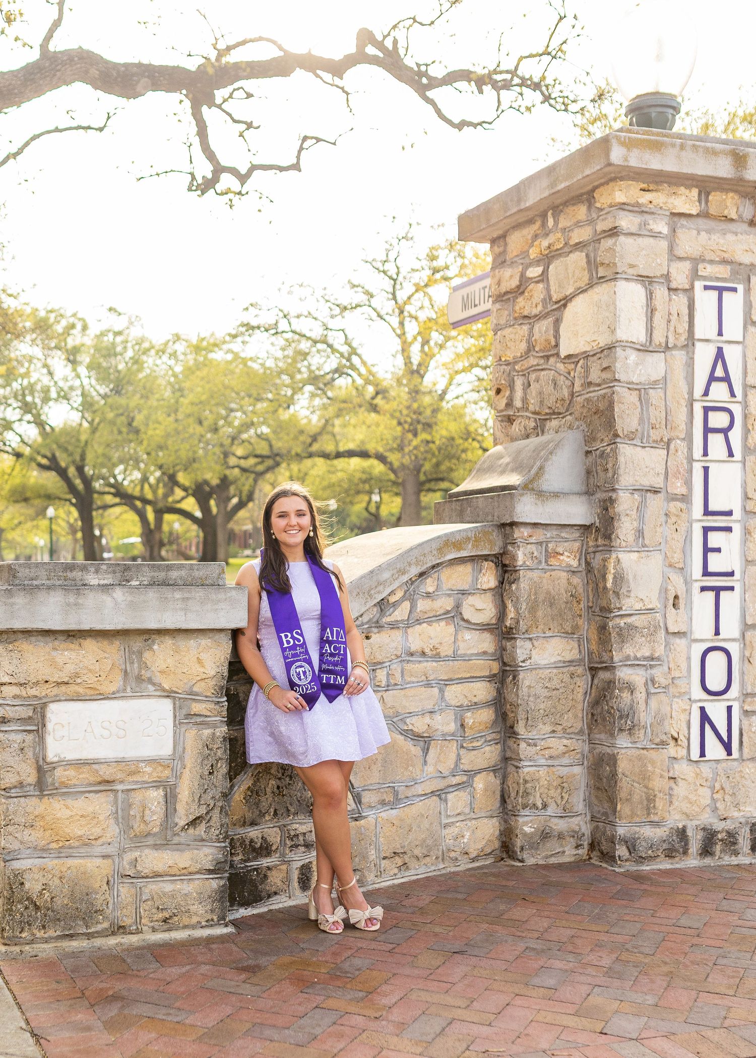 Smiling female graduate in white dress and purple stole posing at Tarleton State University stone entrance gate.