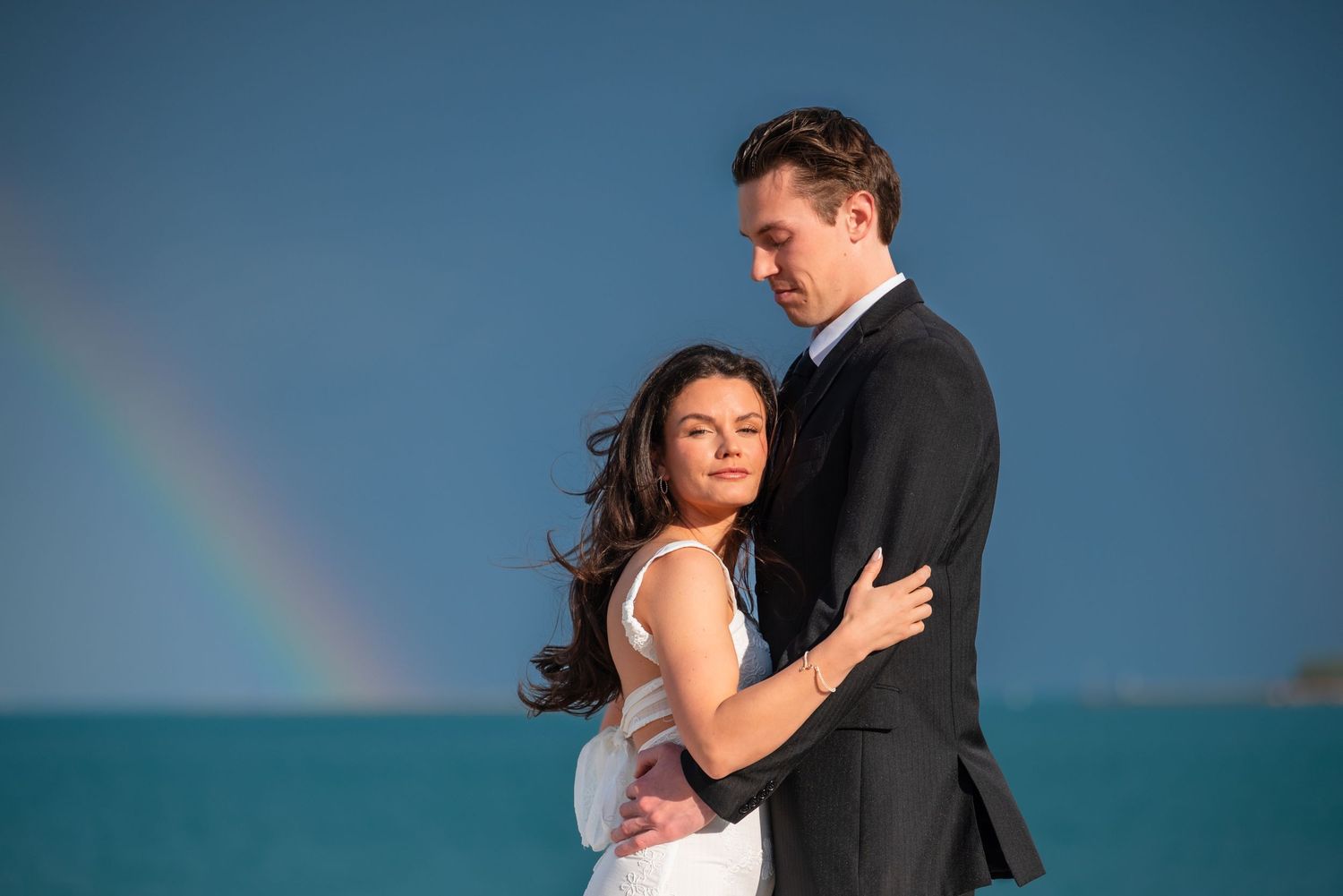 Couple embracing on a beach with a rainbow over lake michigan chicago, bride in white gown and groom in black suit.