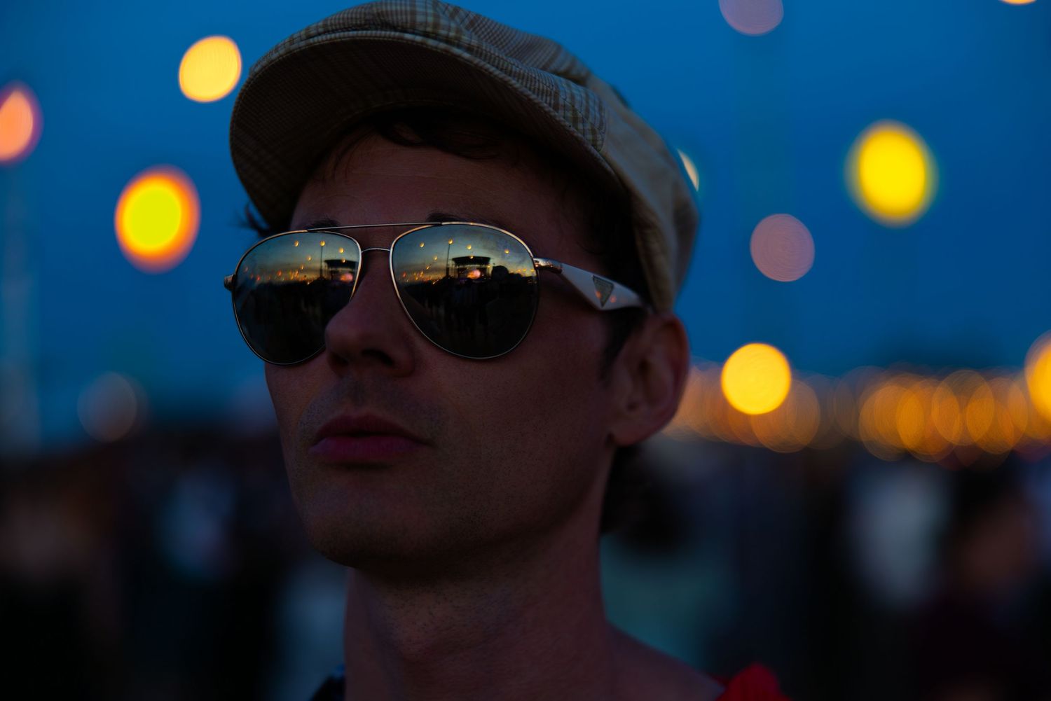 Man wearing cap and aviator sunglasses reflecting city lights at dusk.