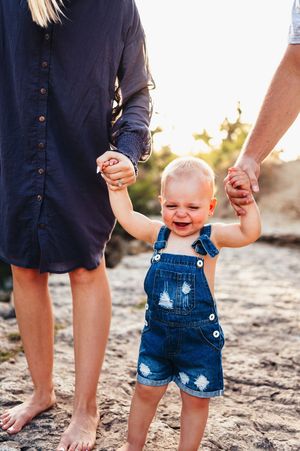Toddler in denim overalls holding parents hands while standing on sandy rocks at sunset.