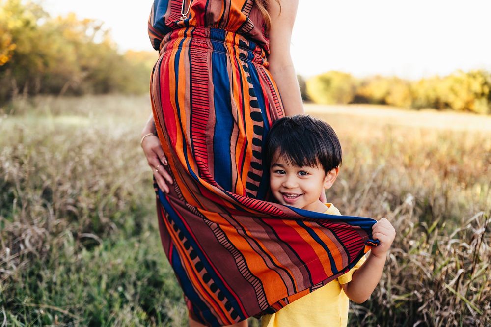 A smiling boy peeks out from behind his pregnant mother's vibrant striped dress in a sunlit meadow.