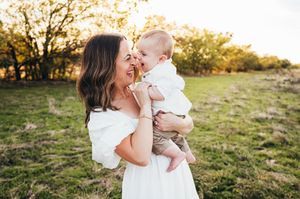 Joyful mother in white dress laughing with her happy baby in a sunny green field.