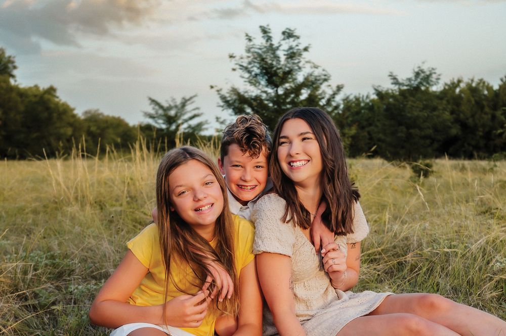 Three siblings sitting together laughing in a sunlit grassy field during a family photo session.
