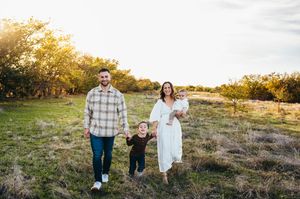 Happy family walking through a sunlit meadow, parents holding toddler's hands in golden hour light.