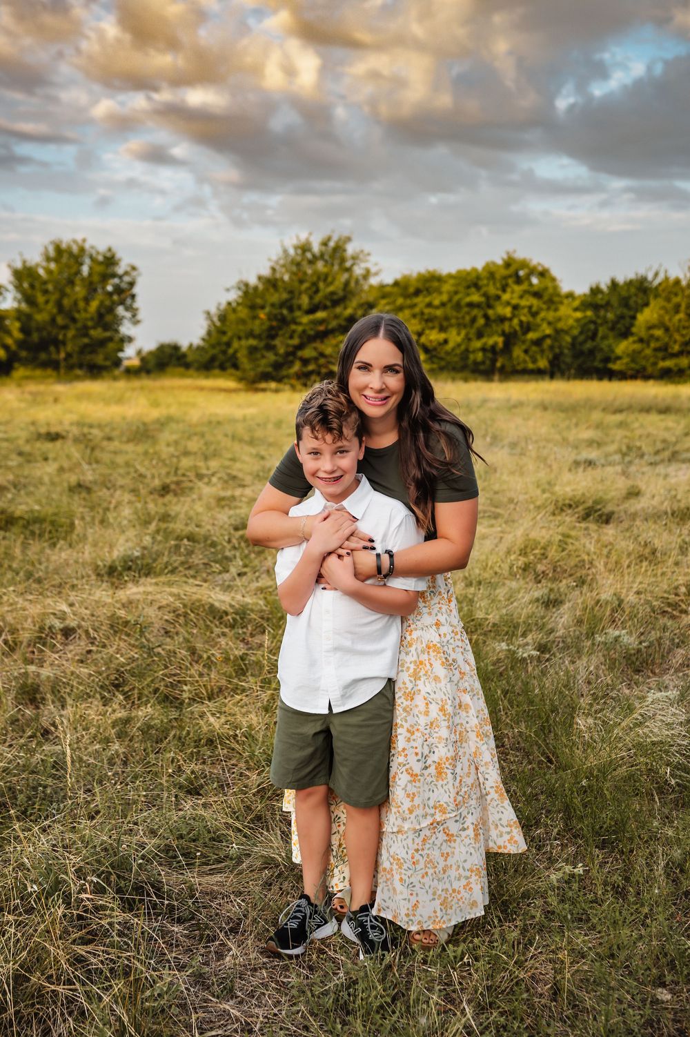 Mother hugging her young son from behind while posing in a green meadow at sunset.