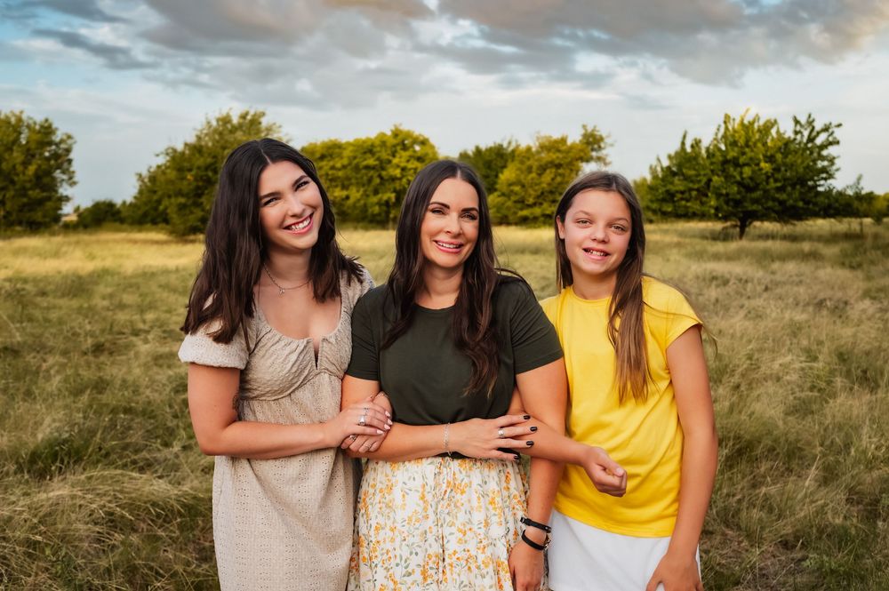 Three smiling brunette women pose together in a golden grassy field at sunset.