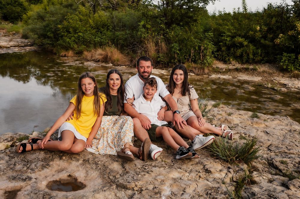 A happy family of five sits together on rocks near a serene river surrounded by lush greenery.