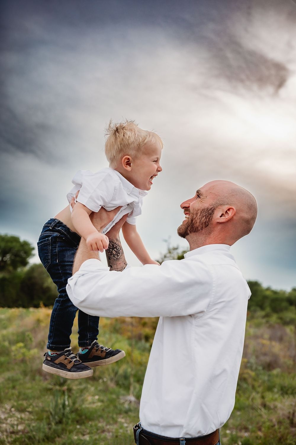 A bald father lifts his laughing blonde toddler son high in the air in an open field.