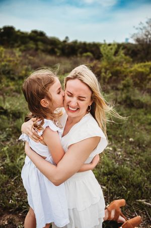 A little girl kisses her laughing blonde mother in a white dress in a green outdoor field.