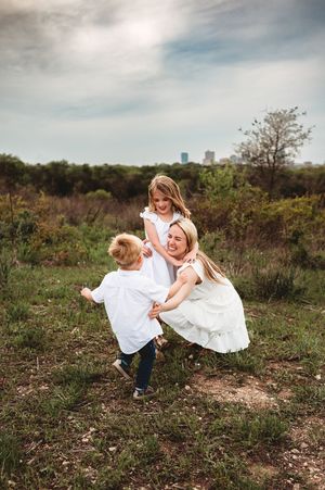 A laughing mother in white dress plays with her two young children in a lush green field.