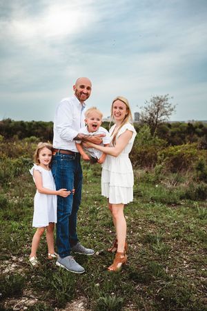 A smiling family of four poses in white outfits in a green field under a cloudy sky.