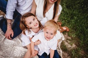 Two happy siblings in white shirts smile while sitting on a lace blanket in a grassy yard.