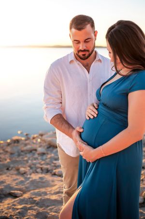 Man tenderly gazing at pregnant partner's belly during golden hour maternity shoot on rocky shore.