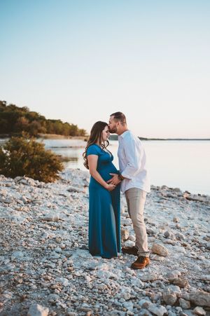 A couple shares a tender kiss while cradling a baby bump on a rocky lakeshore at sunset.