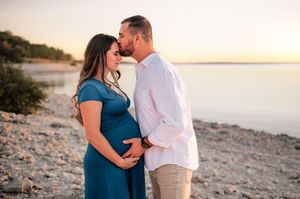 Expecting couple sharing a tender moment at sunset on a rocky beach, woman in teal maternity gown.