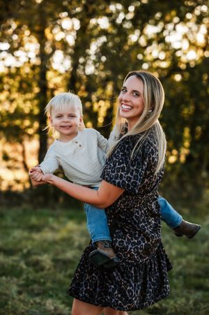 A smiling blonde mother in leopard print dress holds her toddler son in a sunlit outdoor setting.