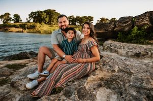 A family of three poses on rocky lakeside terrain, mother in a striped maternity dress at sunset.