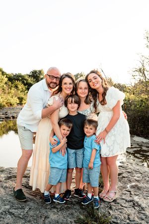 Large family of seven posing together on rocky ground near a pond at golden hour.