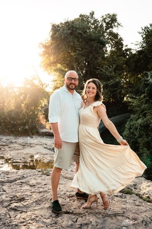 Smiling couple posing together on rocky terrain with golden sunlight streaming through trees behind them.