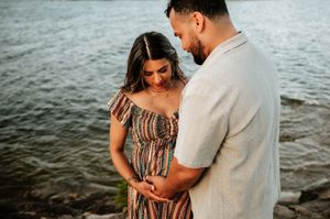A couple lovingly cradles a baby bump together near a shimmering waterfront at golden hour.
