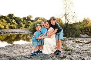 A laughing mom hugs her three young boys in blue on rocky terrain by a river at golden hour.