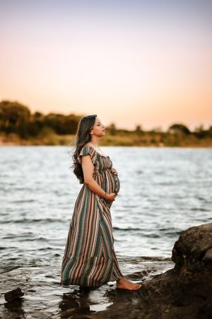 A pregnant woman in a striped maxi dress stands barefoot by the water during a vibrant sunset.