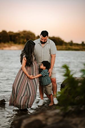 Family maternity photo at sunset by a river, parents and toddler son standing together at the water's edge.
