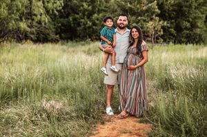 A family of three stands on a dirt path surrounded by lush green grass, mother in striped dress.