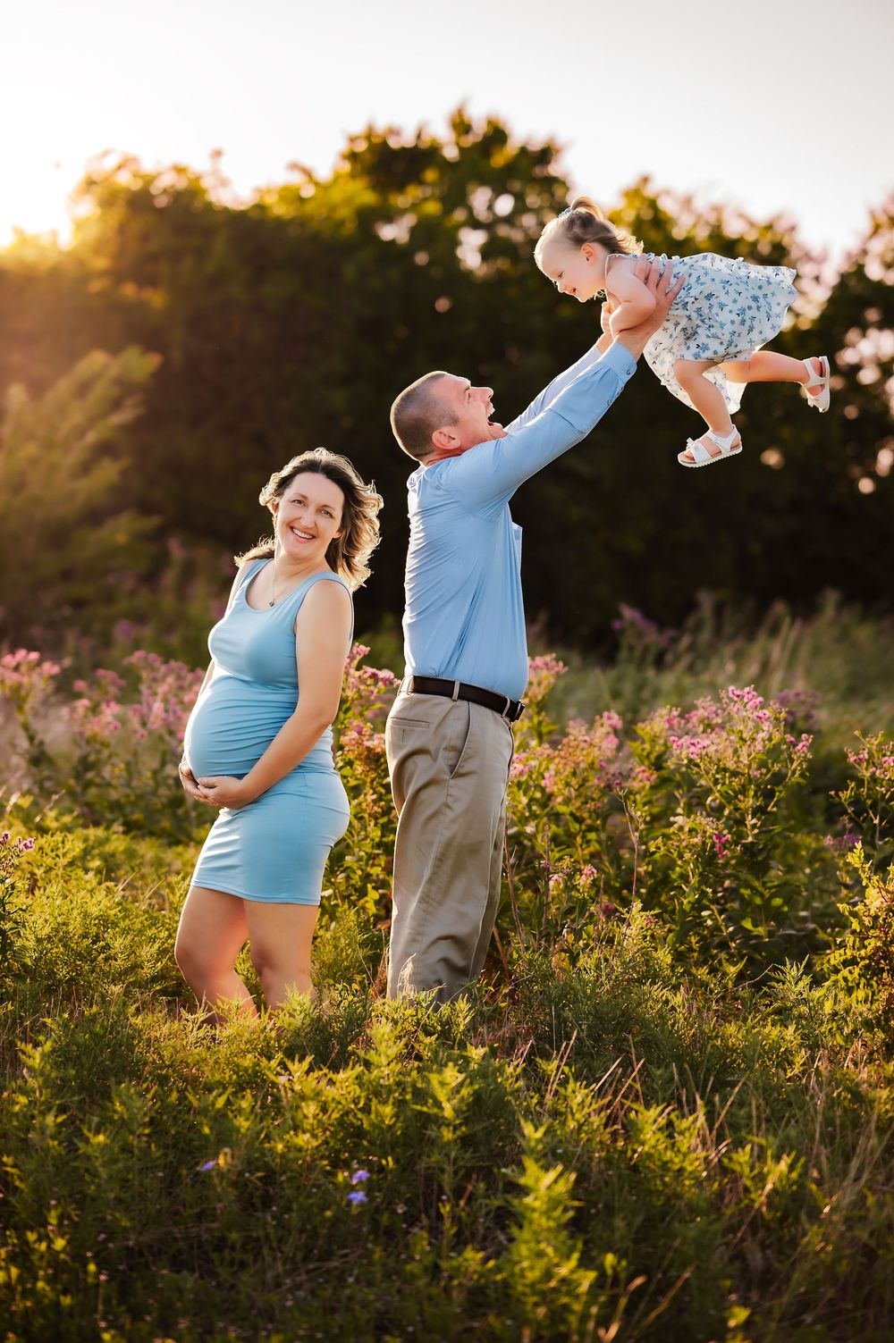 Father lifting laughing toddler girl in wildflower field at sunset beside pregnant mother in blue dress.
