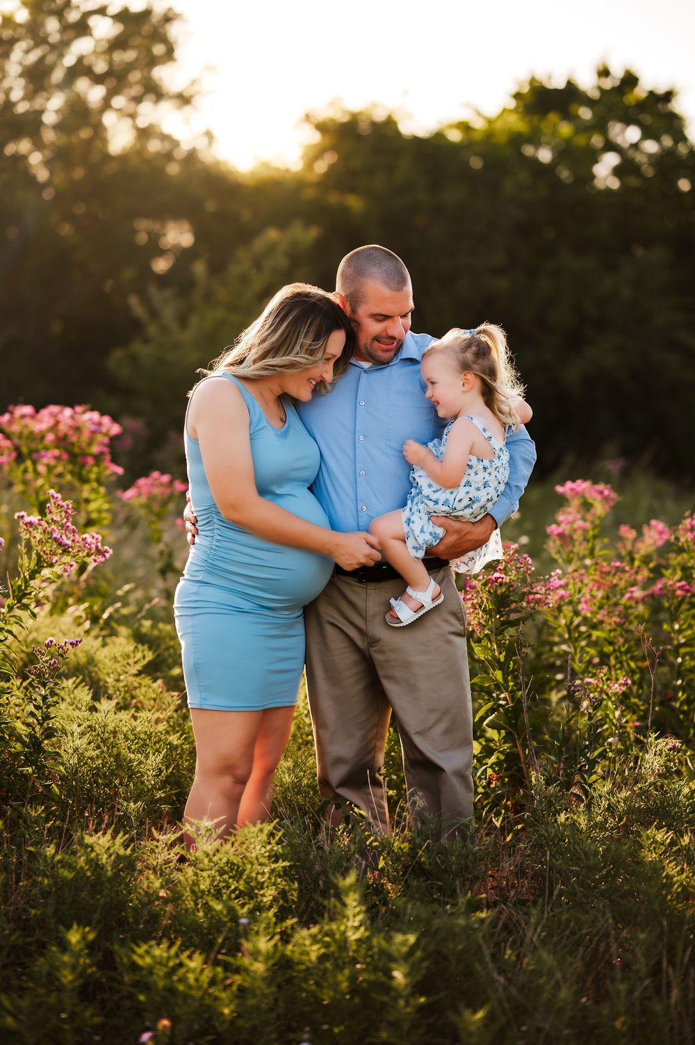 Maternity family portrait in blooming wildflower field at sunset, father holding young daughter close.