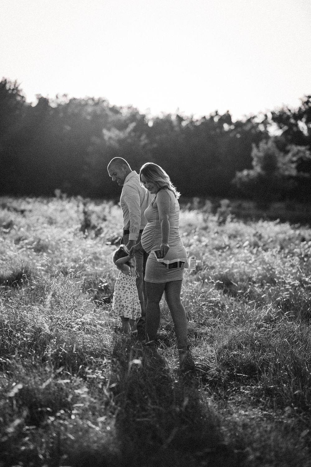 Black and white maternity family photo in sunlit field, couple with toddler daughter at golden hour.