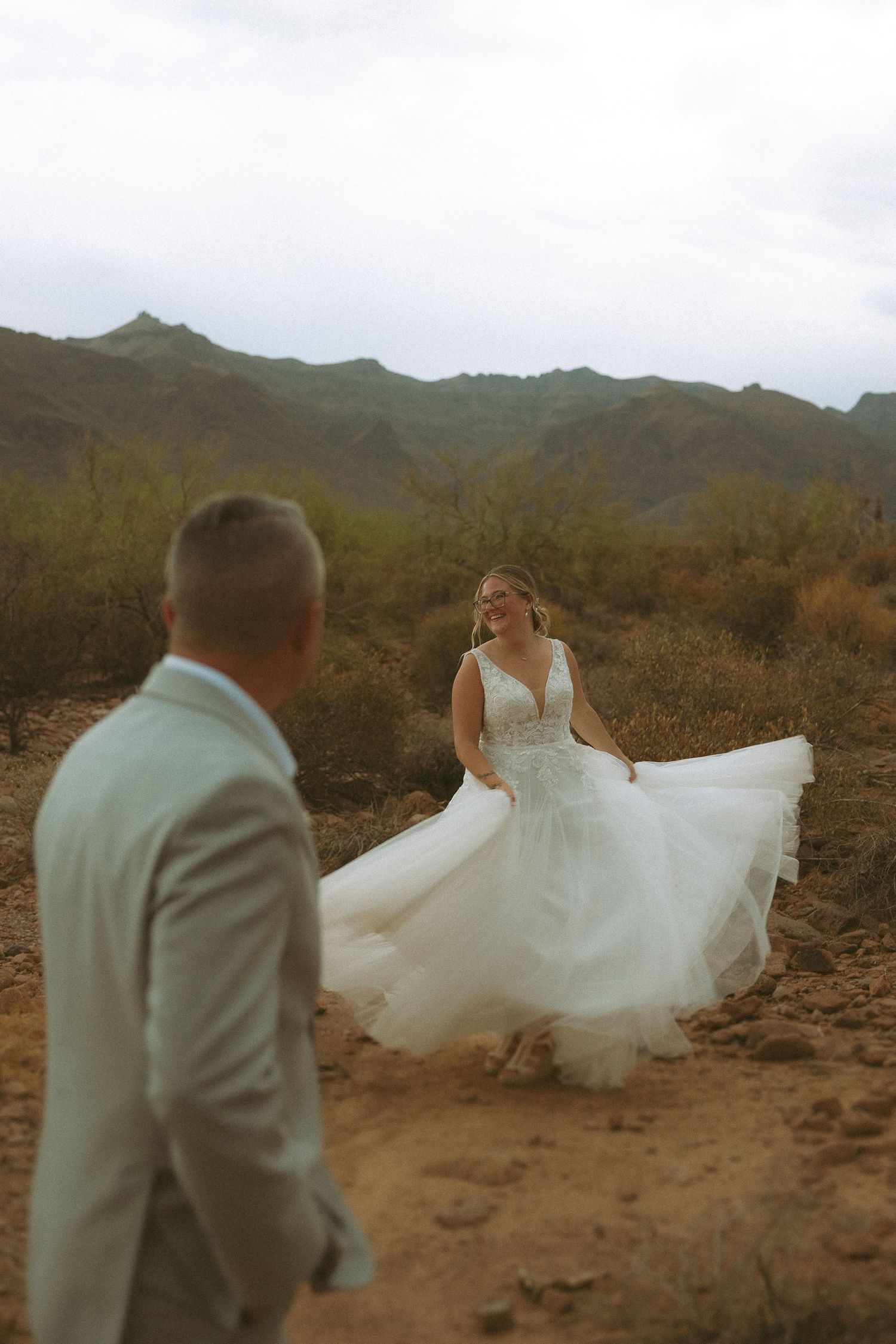 Bride in white tulle gown twirling in desert landscape with mountains, smiling at groom in light suit.