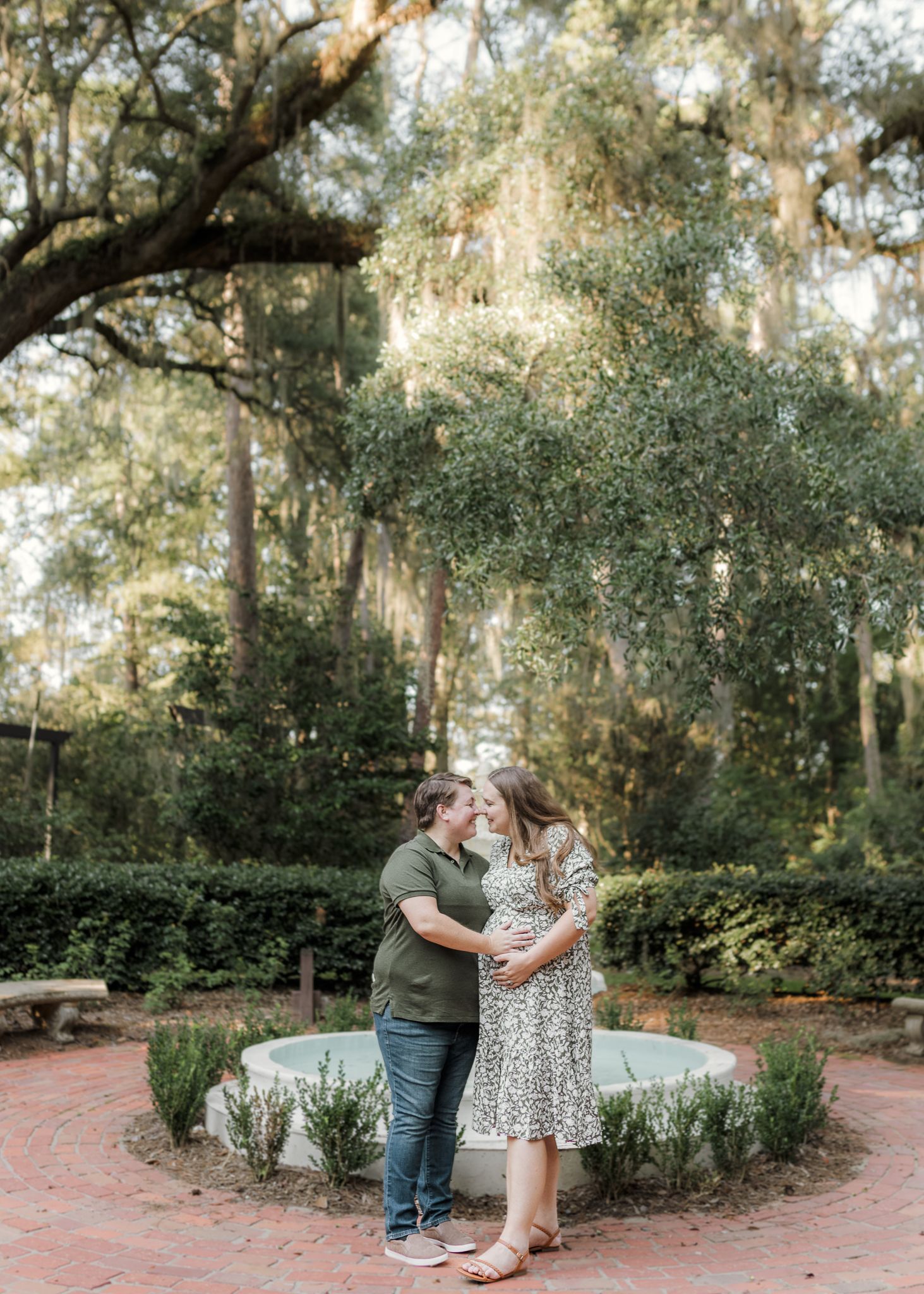 Couple embracing near a garden fountain surrounded by lush trees with Spanish moss during an outdoor maternity photo session.