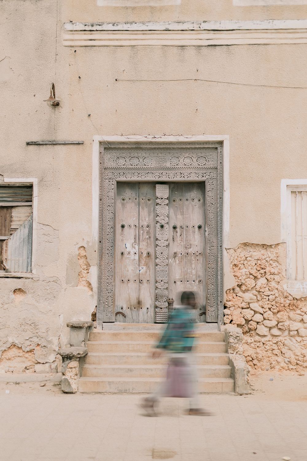 A child walks up stone steps toward an ornate carved wooden door on a weathered sandy building.