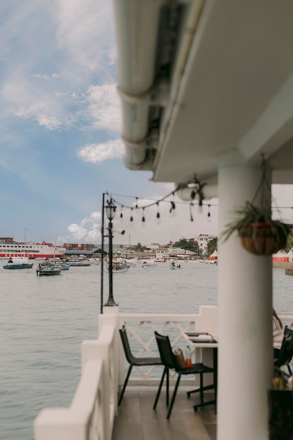 Waterfront restaurant terrace with string lights, chairs, and a scenic harbor view with boats and buildings.
