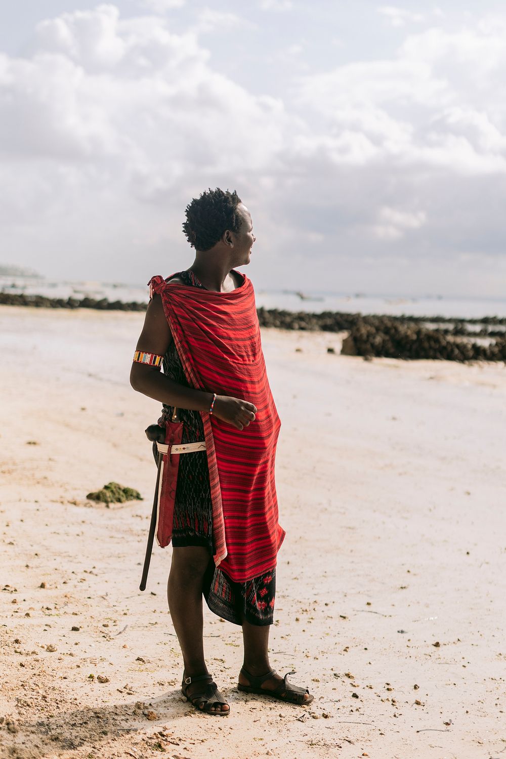 A Maasai warrior in traditional red shuka stands on a sandy beach, holding a wooden staff, with cloudy skies behind.