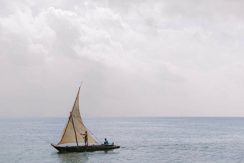 Two people sailing a traditional wooden dhow with a tan sail on calm ocean waters under a cloudy sky.